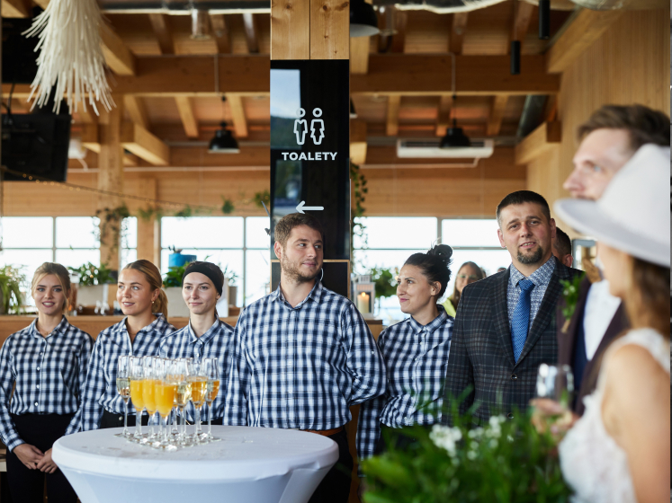 Obsługa w szachowniczych koszulach stoi wokół stołu z kieliszkami szampana, obserwując pannę młodą w białym kapeluszu w drewnianym wnętrzu restauracji